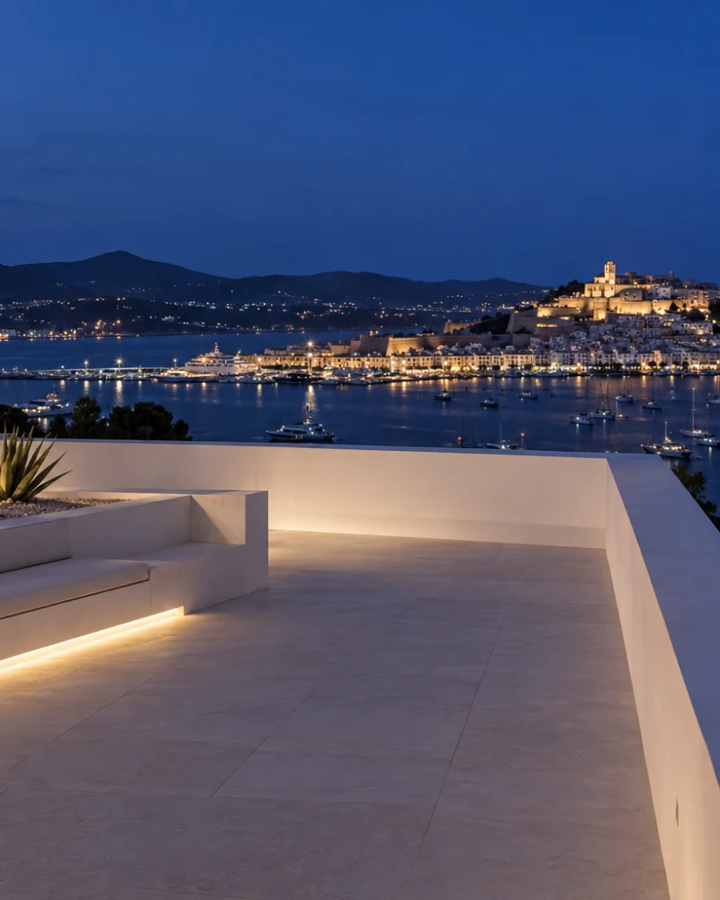 White roof terrace at night with lit built-in bench, agaves and a view over the bay to Dalt Vila